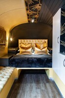 A bedroom with a tufted headboard, two gold pillows, a marble-like bedspread, and a wooden floor.