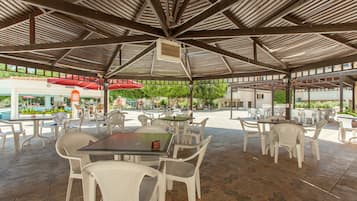 An outdoor patio area with tables and chairs, a covered seating area, and a view of the pool and greenery in the background.