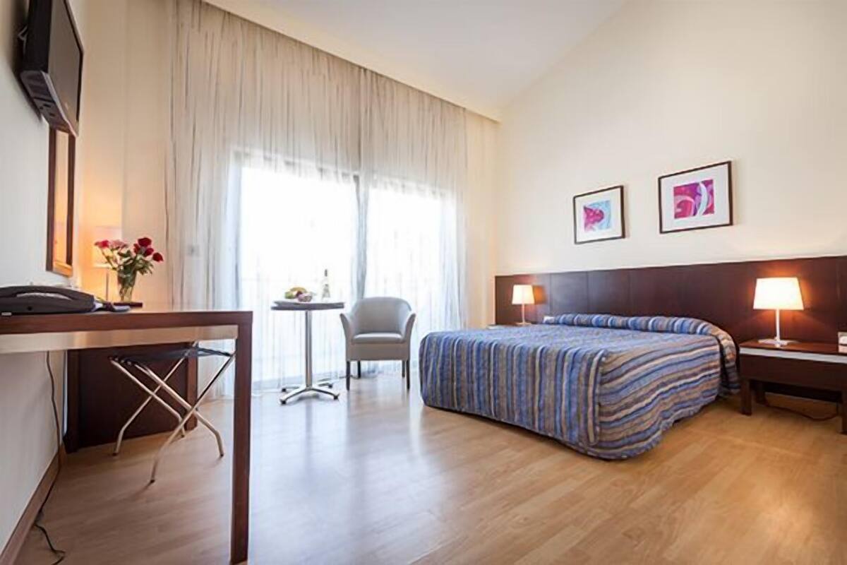 Une chambre d’hôtel comprenant un lit, un bureau avec un téléphone, une chaise, une petite table avec une lampe, et deux tableaux encadrés accrochés au mur.
