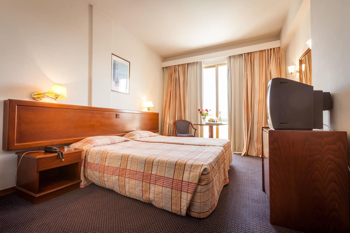 Une chambre d’hôtel avec un grand lit, un bureau avec un téléphone, un téléviseur posé sur une commode en bois et une fenêtre avec des rideaux.
