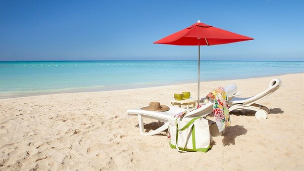 Escena de playa con dos sillas blancas, un paraguas rojo, un sombrero, un bolso y una pequeña mesa con una bebida.