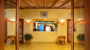 Un área de recepción con techo de madera, paredes amarillas y un mostrador de recepción. Hay dos recepcionistas, plantas en macetas y bancos de madera.