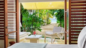 Una habitación con cama, una mesita con un florero, una silla y vistas a un jardín tropical y una playa.