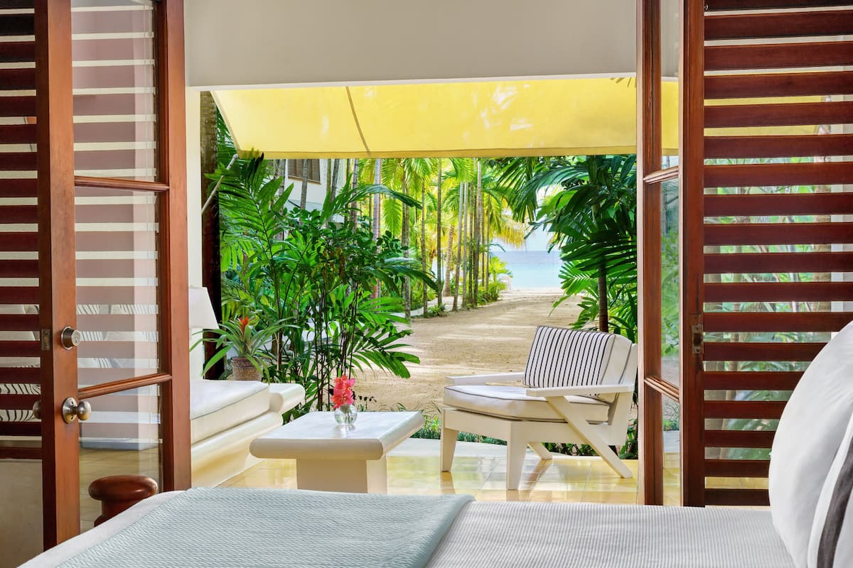 A room with a bed, a small table with a vase, a chair, and a view of a tropical garden and beach.
