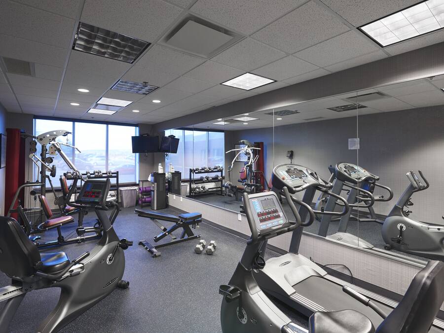 Un gimnasio con diverso equipo de ejercicio, incluyendo cintas de correr y máquinas de pesas, y amplias ventanas que permiten la entrada de luz natural.