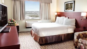 A hotel room with a large bed, a desk with a TV, a window with curtains, a framed picture on the wall, and a patterned rug. - Capital Hotel (St. John's)