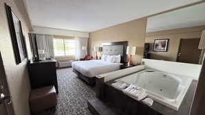 A hotel room with a large bed, a smaller bed, a desk, a chair, a bathroom with a bathtub, and a window with curtains. - Grand Oaks Hotel (Branson)