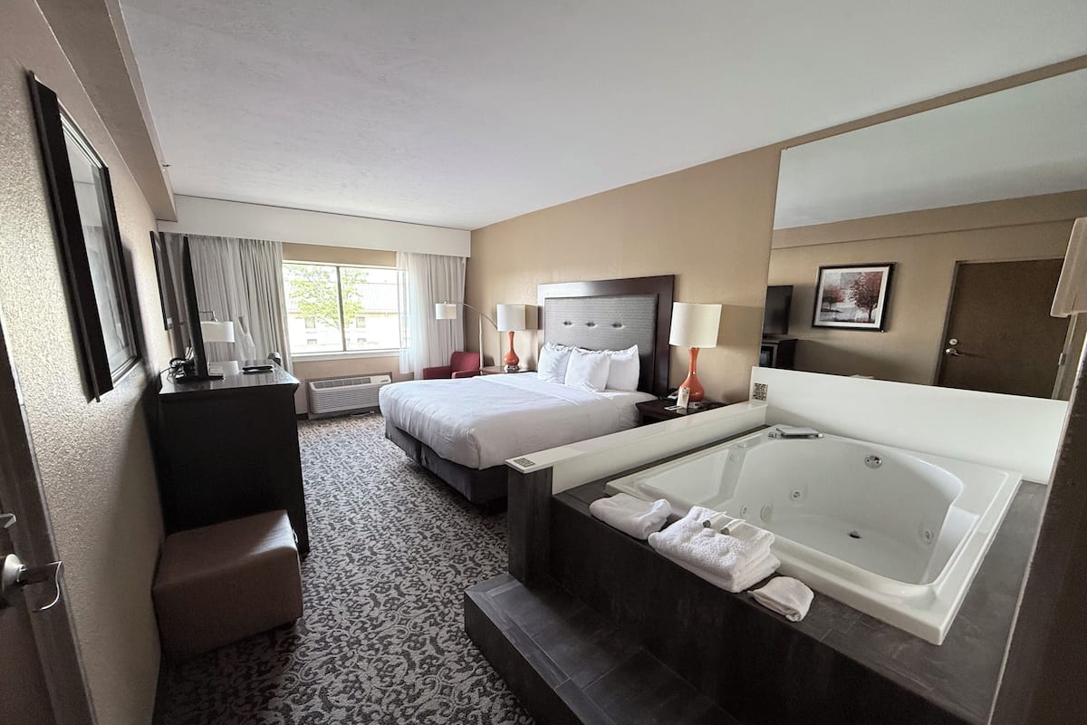A hotel room with a large bed, a smaller bed, a desk, a chair, a bathroom with a bathtub, and a window with curtains.