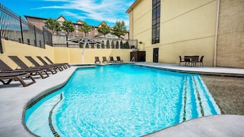 A swimming pool with lounge chairs and a table, surrounded by a building with a brick facade and a fence.