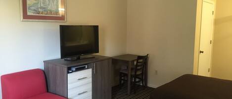 Quarto de hotel com televisão sobre um móvel de madeira, uma cadeira vermelha, uma mesa e uma cama.