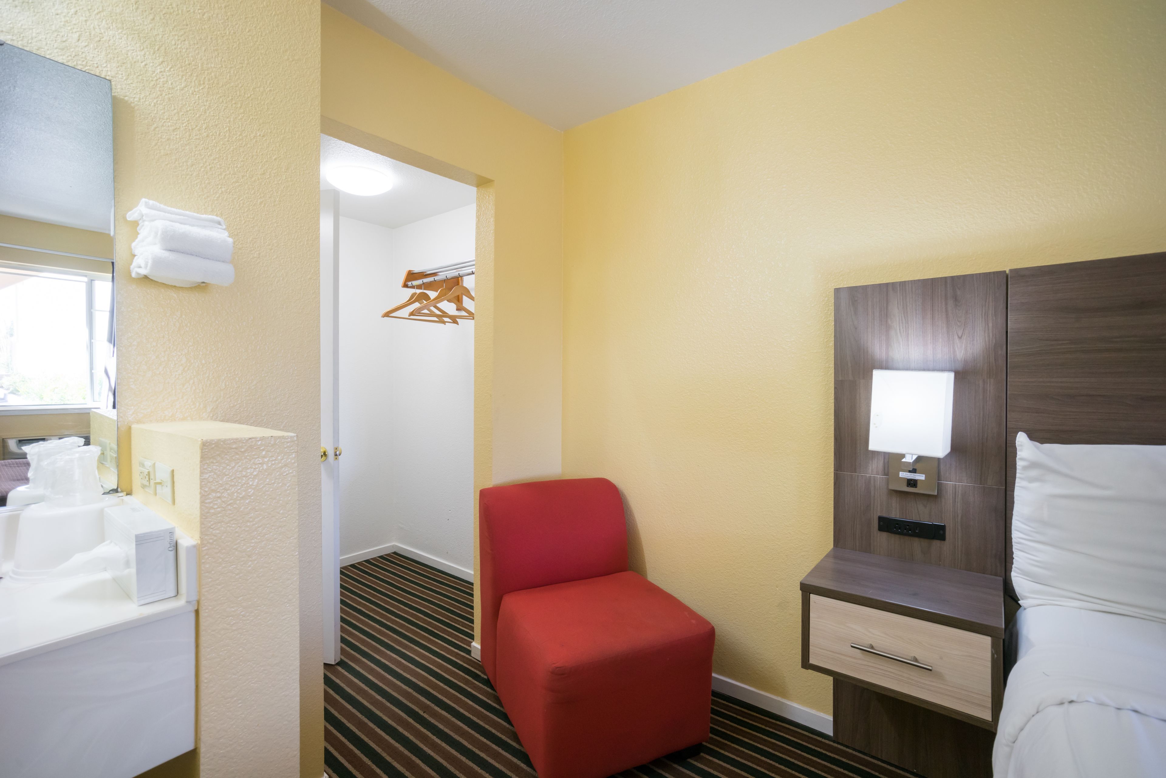 Quarto de hotel com cama, mesa de cabeceira, uma cadeira vermelha, banheiro com pia e espelho, e corredor com porta-roupas.