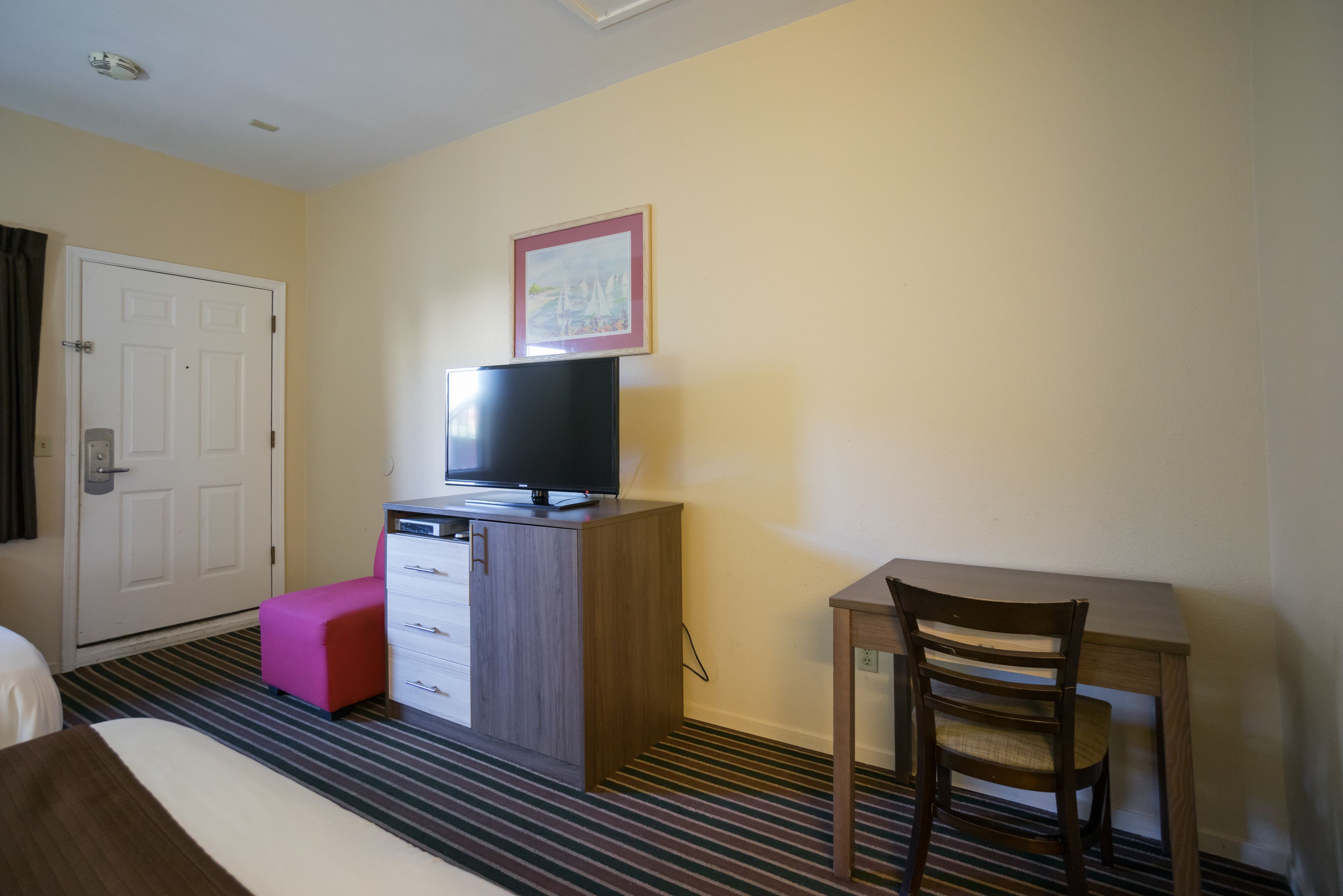 A hotel room with a bed, a desk, a television on a cabinet, a pink ottoman, and a framed picture on the wall.