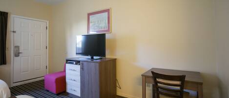 Une chambre d’hôtel avec un lit, un bureau, un téléviseur posé sur un meuble, un pouf rose et un tableau encadré accroché au mur.