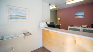 Un área de recepción con mostrador, escritorio y una silla. Hay cuadros enmarcados en la pared y un estante pequeño con revistas.