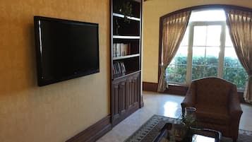A living room with a flat-screen TV, a bookshelf, a patterned rug, a glass coffee table, and a view through a large window with curtains.