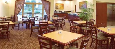 A hotel dining area with multiple tables and chairs, a large window with curtains, a bar area, and a plant.