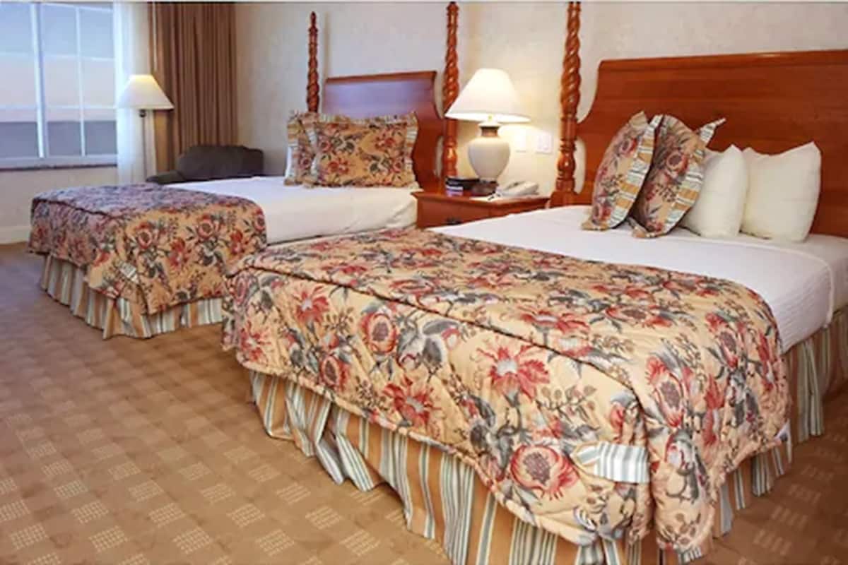 Hotelkamer met twee bedden, een houten hoofdbord, een nachtkastje met een lamp en een raam met gordijnen.