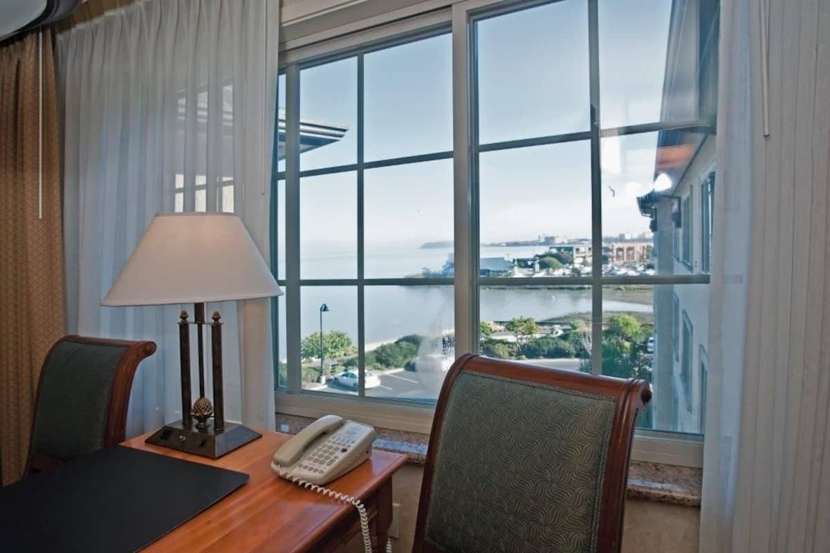 Hotelkamer met groot raam, uitzicht op een waterpartij en gebouwen. Er is een bureau met telefoon en lamp.