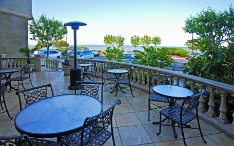 An outdoor patio with round tables and chairs, a view of the sea, and parked cars in the background.