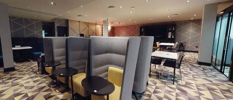 A modern hotel lobby with geometric patterned flooring, seating areas with tables and chairs, and a large flat-screen TV on the wall.