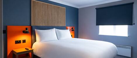 A hotel room with a bed, a nightstand, a wall-mounted lamp, a window with a blind, and a radiator.