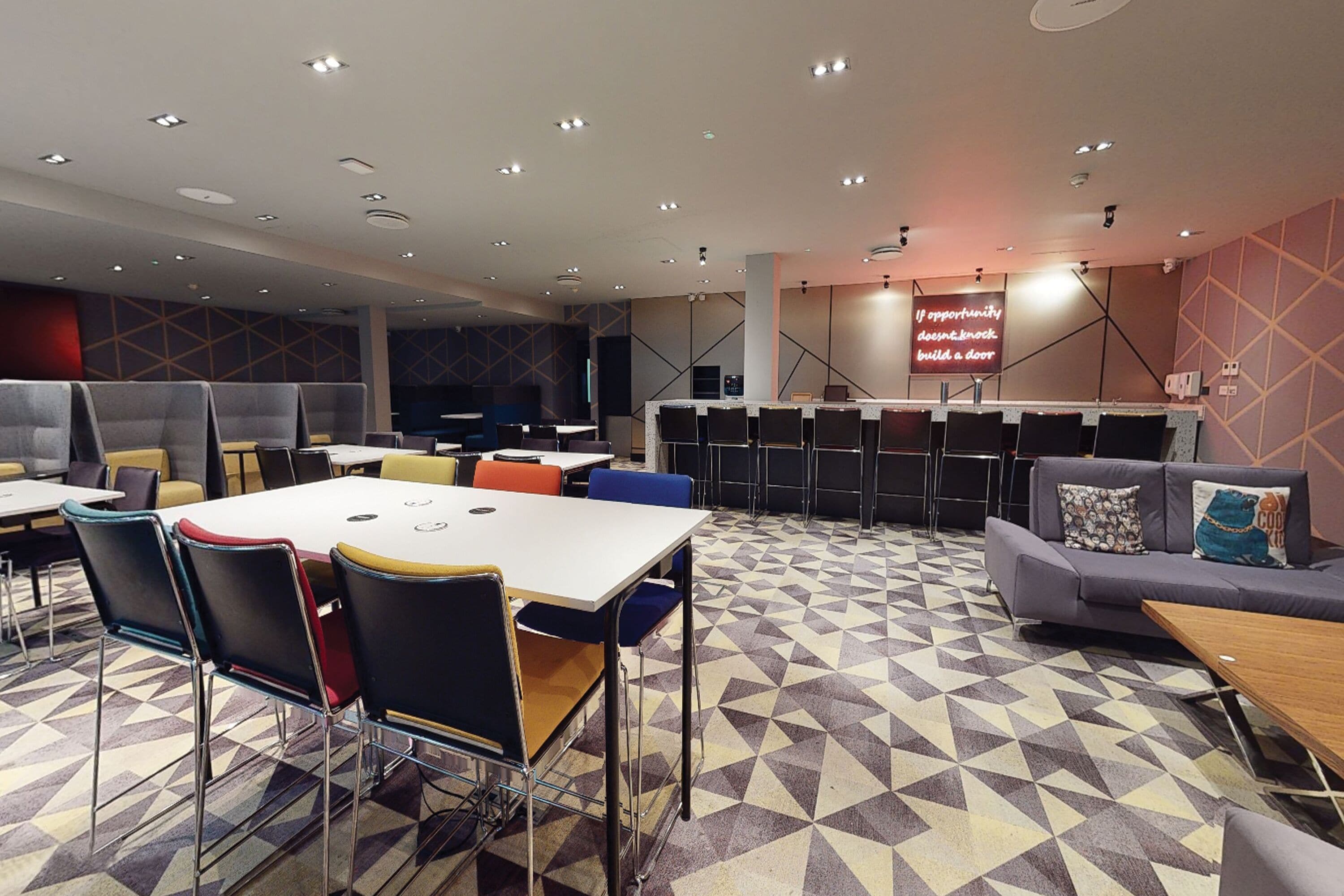 A modern conference room with a geometric patterned floor, multiple tables and chairs, and a large screen displaying motivational text.