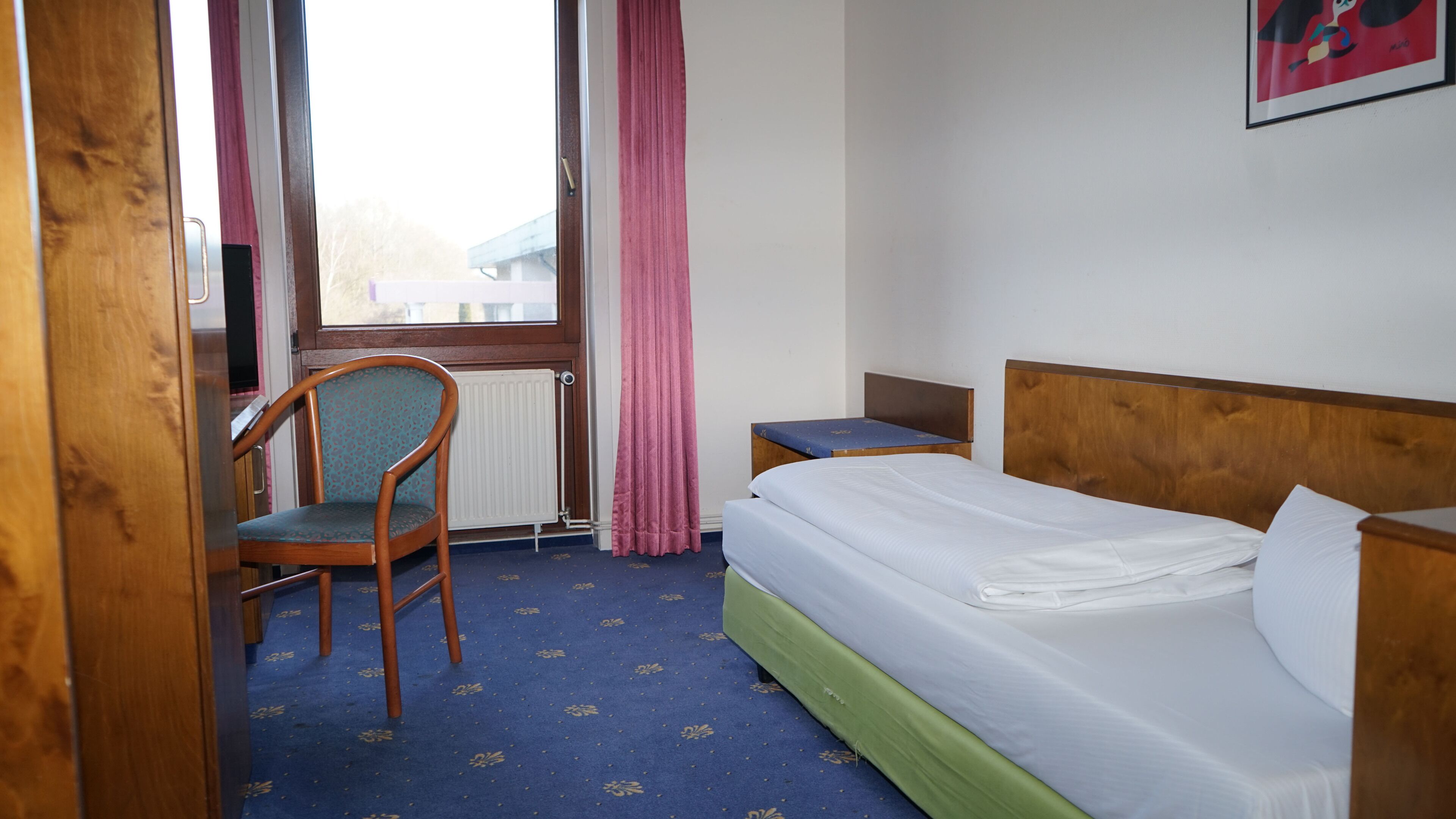 Ein Hotelzimmer mit einem hölzernen Bett, einem Sessel, einem Schreibtisch und einem Fenster mit Vorhängen.