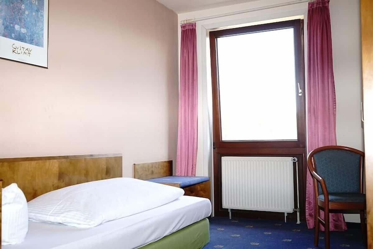 A hotel room with a bed, a chair, a small table, a window with curtains, and a radiator.
