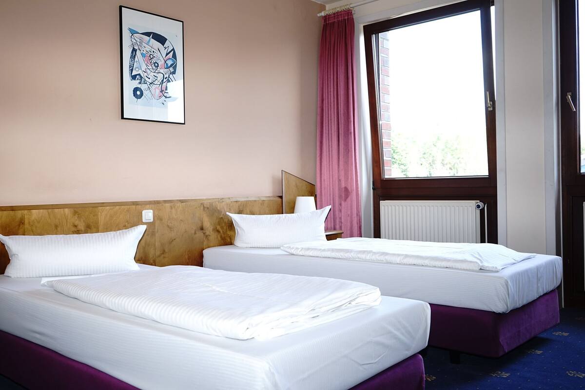 A hotel room with two beds, a wooden headboard, a nightstand with a lamp, a window with a view, and a painting on the wall.
