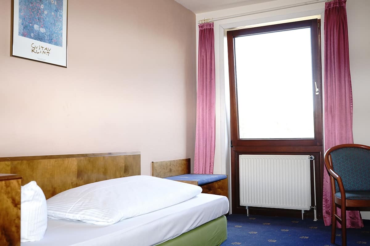 A hotel room with a bed, a chair, a window with curtains, a radiator, and a framed picture on the wall.