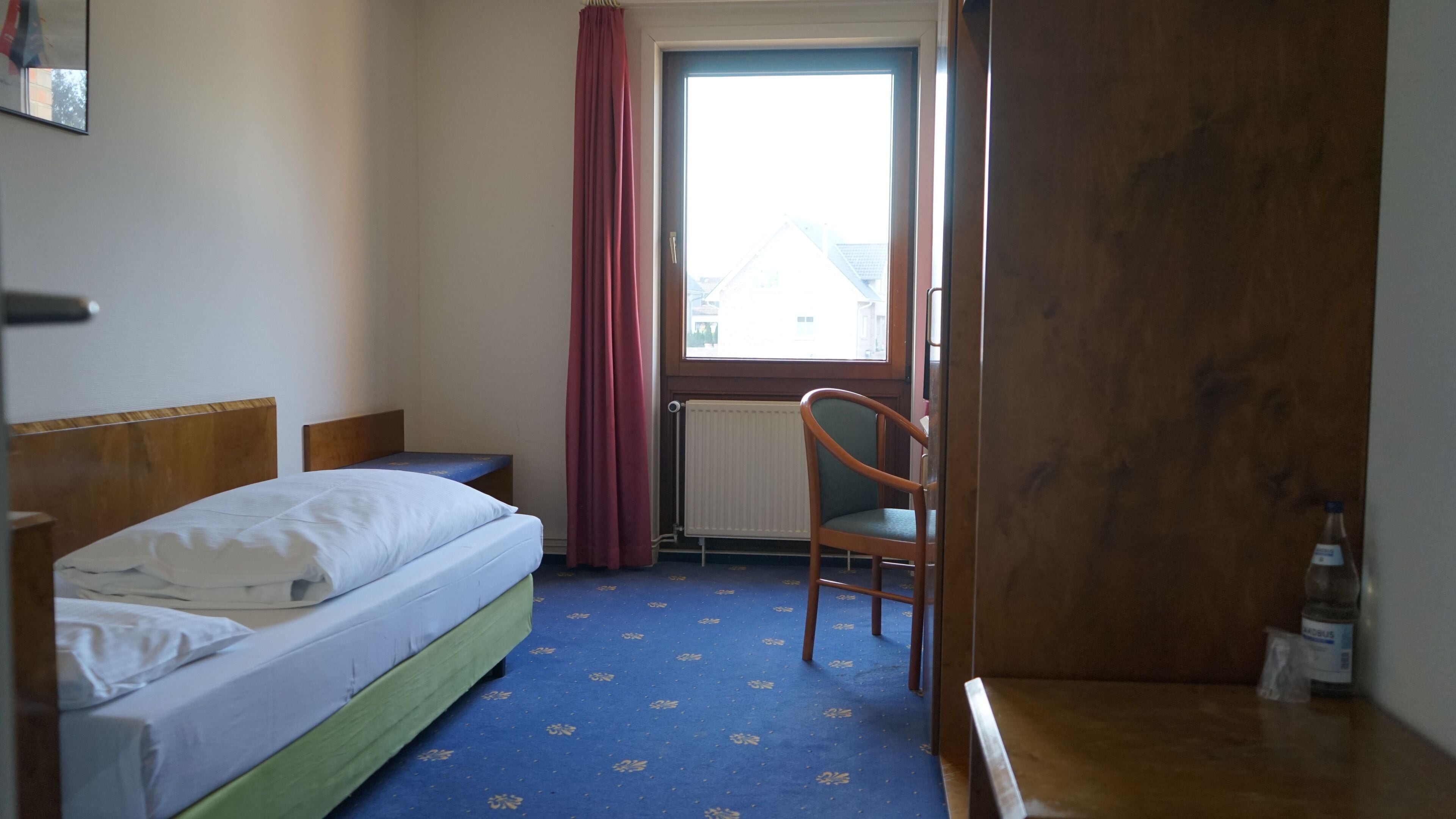 A hotel room with a bed, a chair, a radiator, a window with curtains, and a wooden door.
