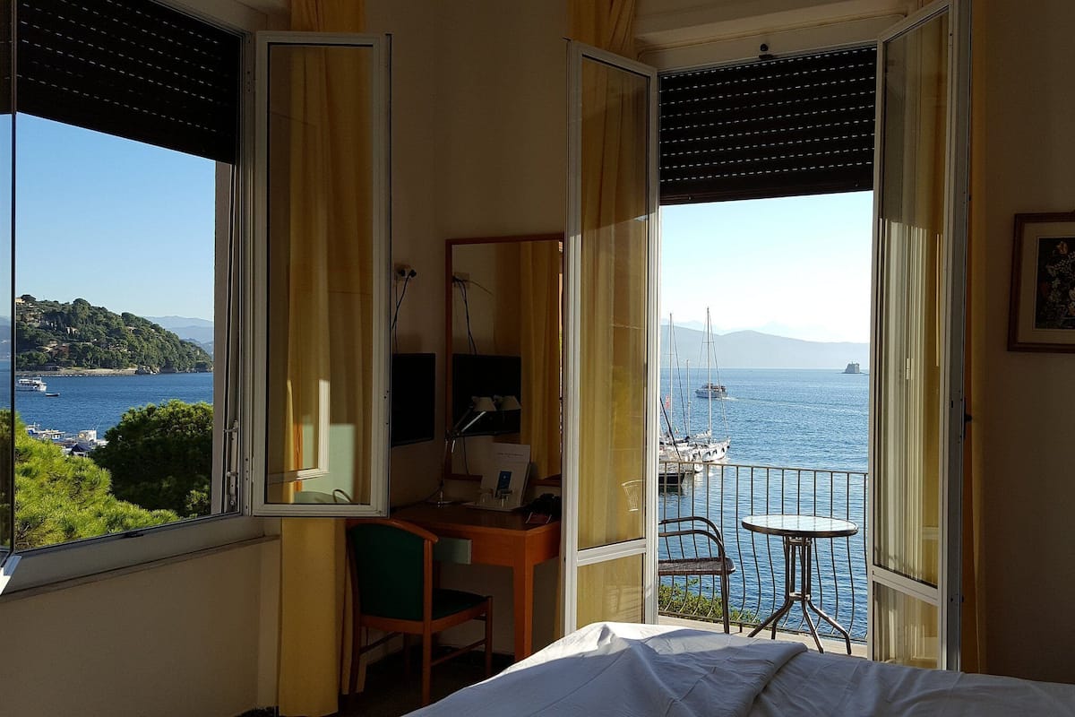 A hotel room with a bed, a desk, a chair, and a small table by a window overlooking the sea.
