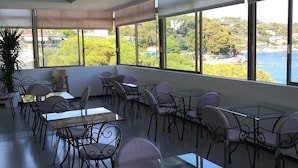 A spacious indoor area with multiple tables and chairs, large windows offering a view of the sea and greenery. - Hotel Belvedere (Portovenere)