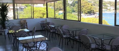 A spacious indoor area with multiple tables and chairs, large windows offering a view of the sea and greenery.