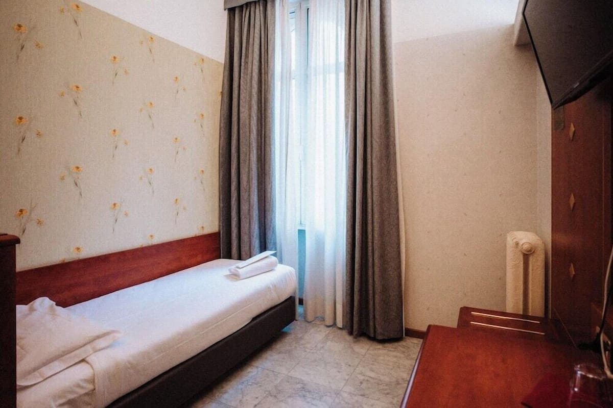 A hotel room with a bed, a desk, a radiator, and a window with curtains.