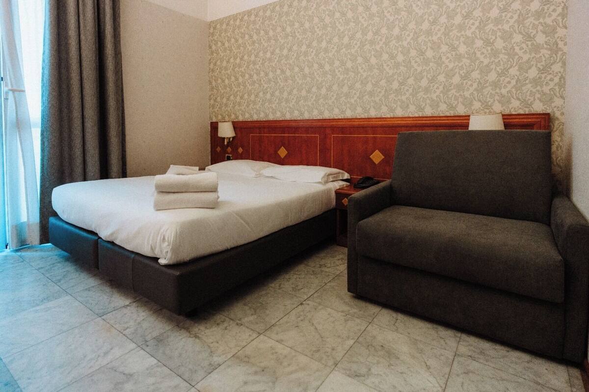 A hotel room with a bed, a chair, a nightstand, and a lamp.