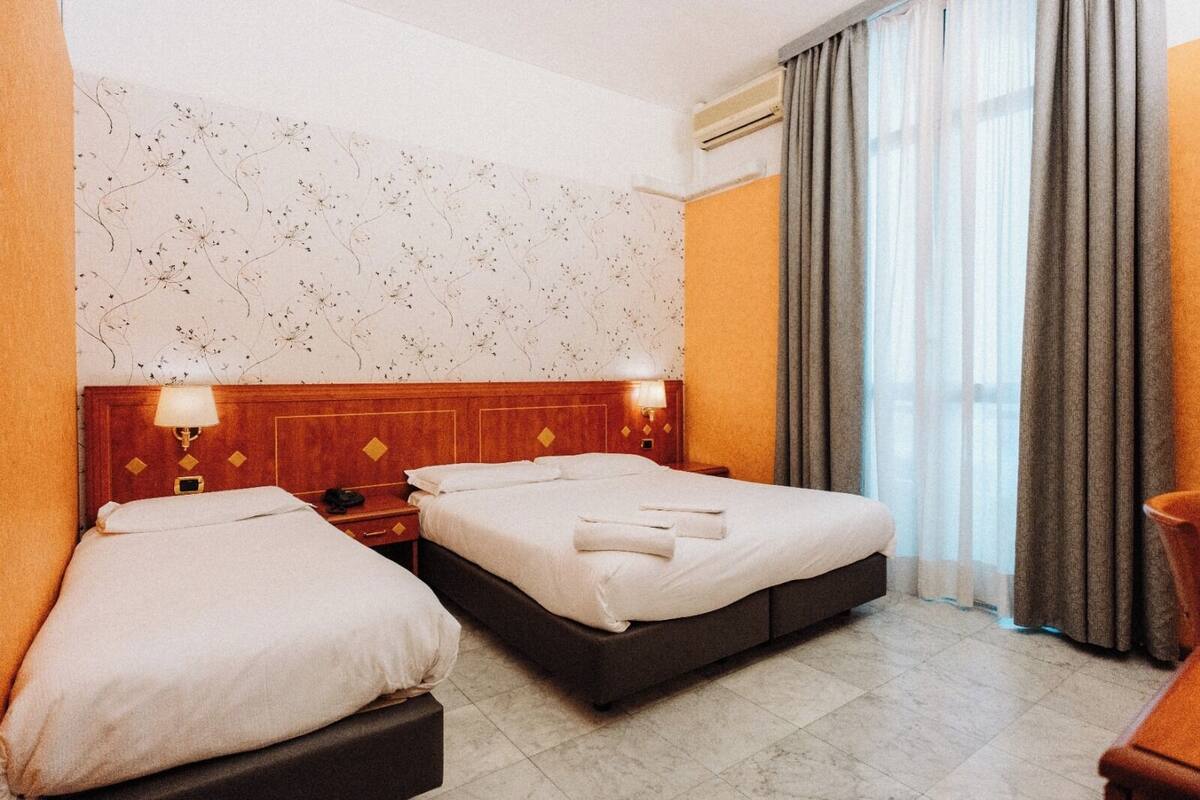 A hotel room with two beds, a wooden headboard, a nightstand, a lamp, and a window with curtains.