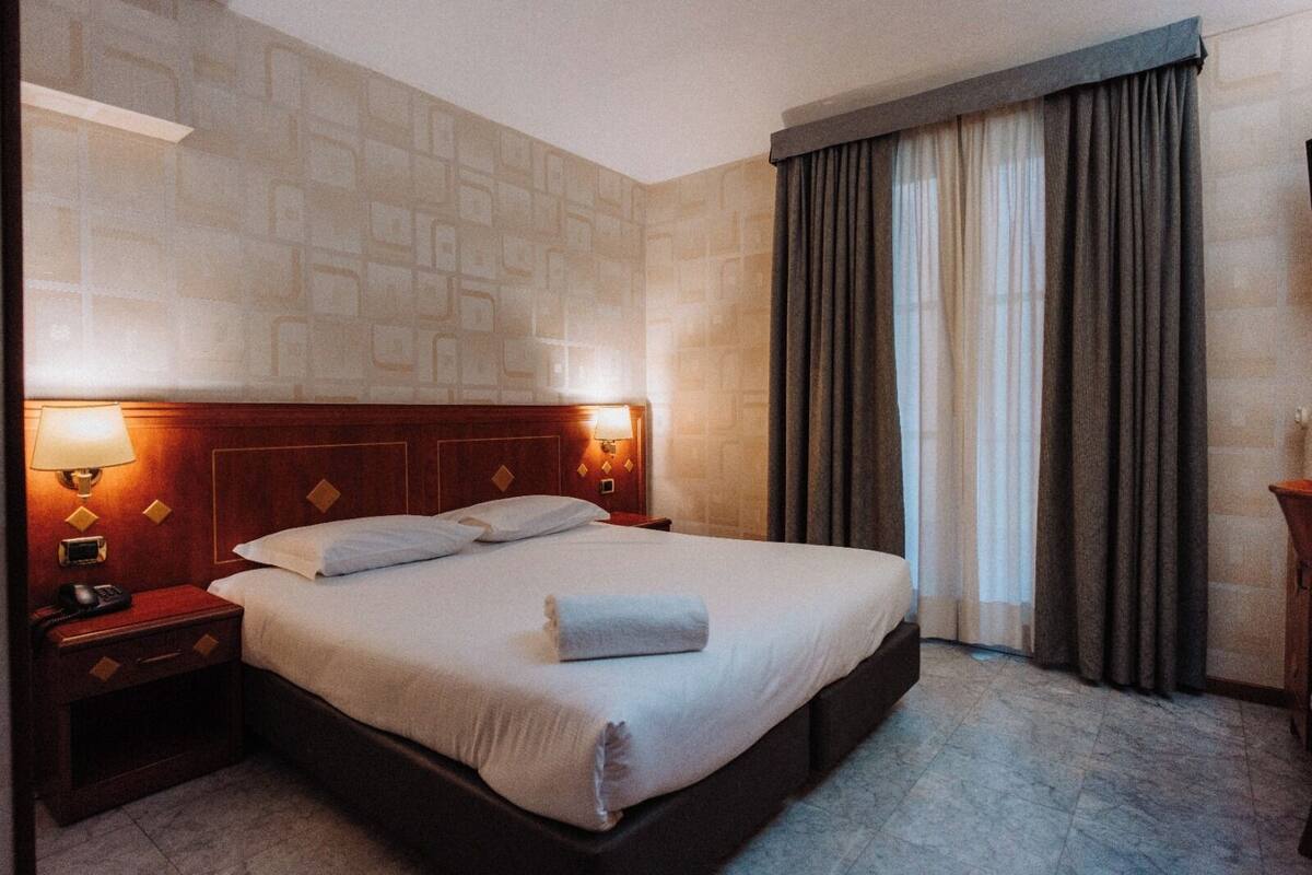 A hotel room with a large bed, a wooden headboard, a nightstand with a lamp, and a window with curtains.