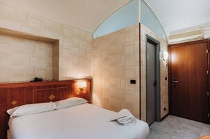 Habitación de hotel con cama, cabecera de madera, mesita de noche con lámpara, puerta y pared con un diseño geométrico.