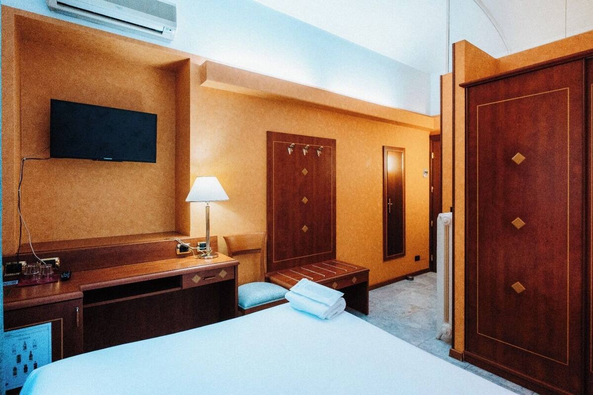 A hotel room with a bed, a desk with a lamp, a TV mounted on the wall, and two wooden doors.