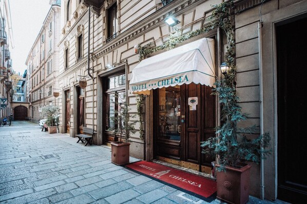 A hotel entrance with a red carpet, a sign reading "CHELSEA HOTEL", and a small tree in a planter. - Hotel Chelsea (Turin)