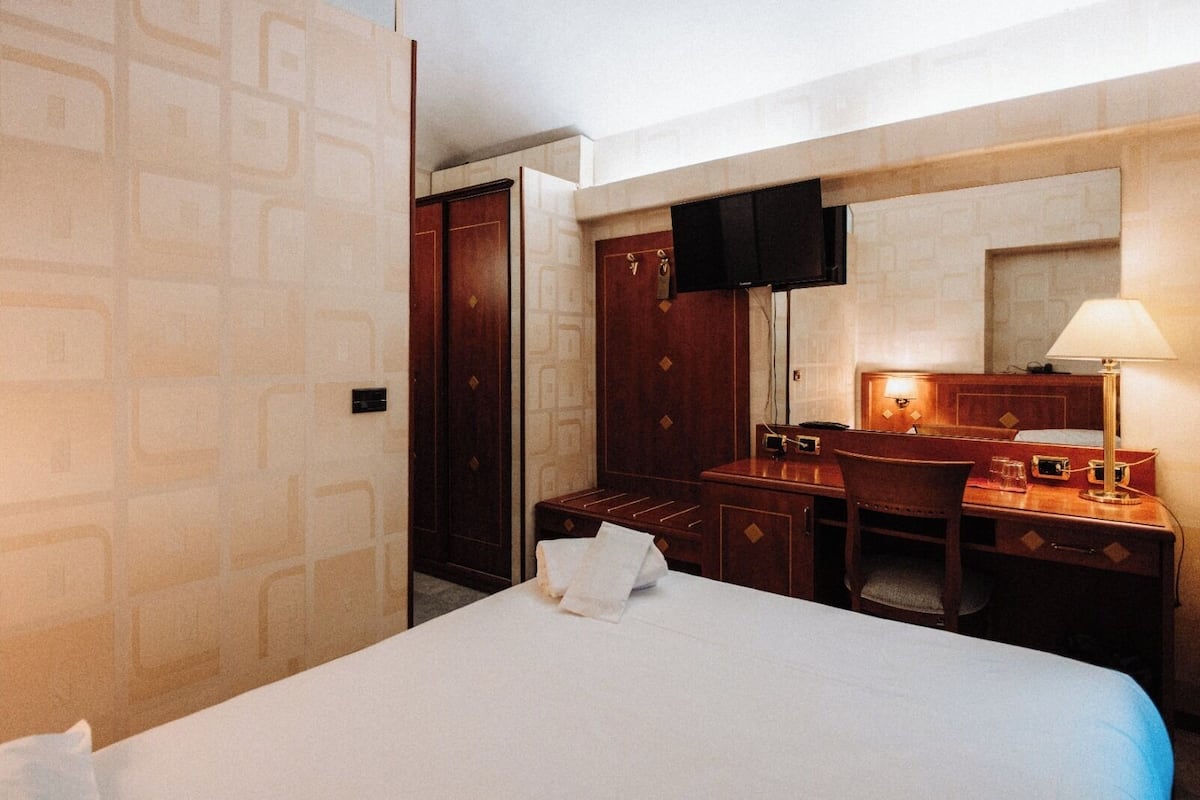 A hotel room with a bed, a desk with a chair, a mirror, a television, and a wardrobe.