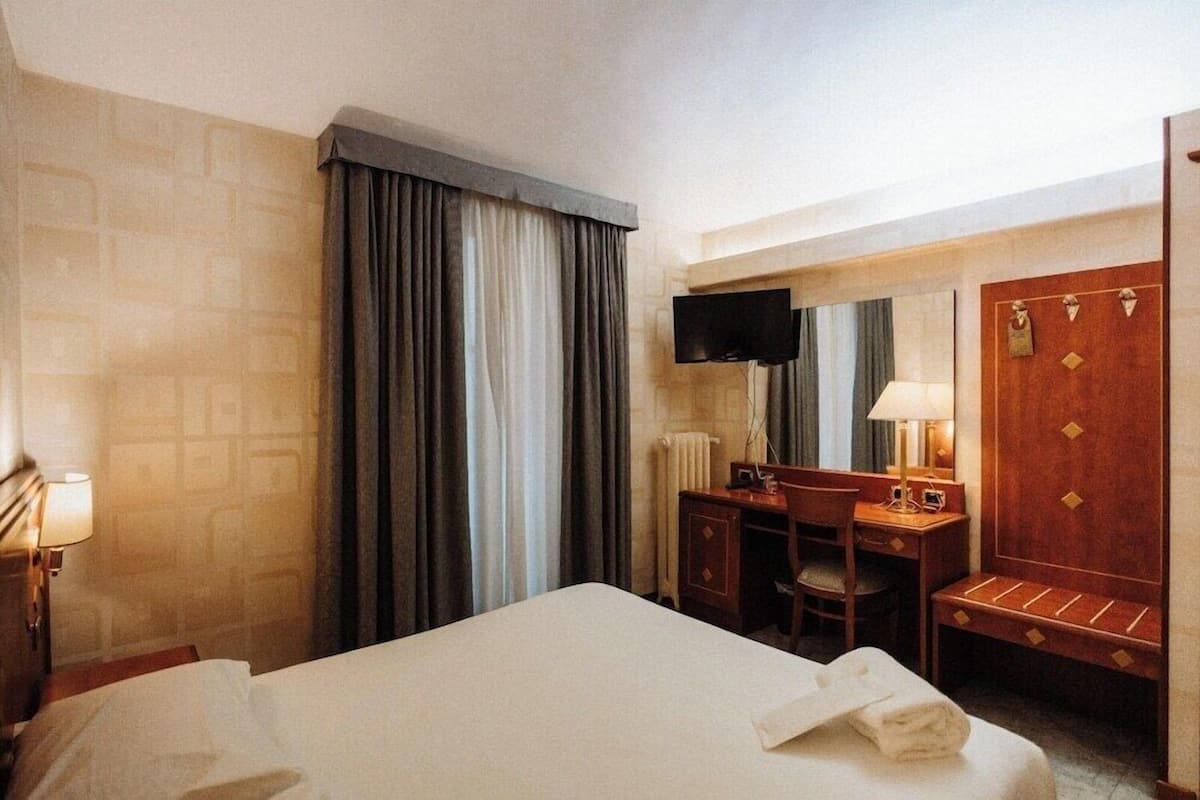 A hotel room with a bed, a desk with a chair, a television, a mirror, and a wardrobe.