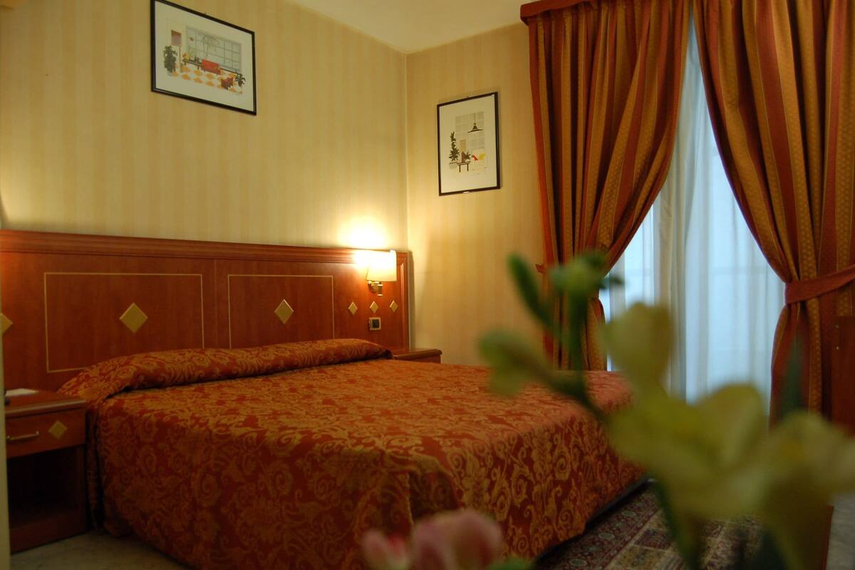 A hotel room with a large bed, a nightstand, a wall-mounted lamp, and two framed pictures on the wall.
