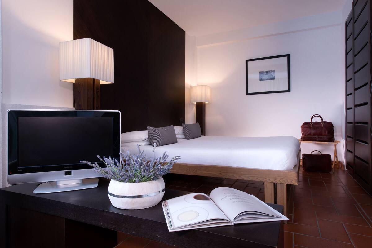 Ein modernes Hotelzimmer mit einem Bett, Nachttischlampen, einem Flachbildfernseher, einem Holznachttisch, einer Topfpflanze und einem offenen Buch.