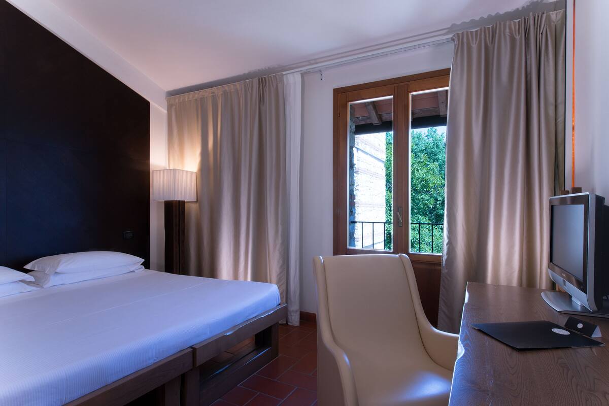 Ein Hotelzimmer mit einem Bett, einem Schreibtisch mit Laptop, einem Sessel, einem Fernseher und einem Fenster mit Vorhängen.