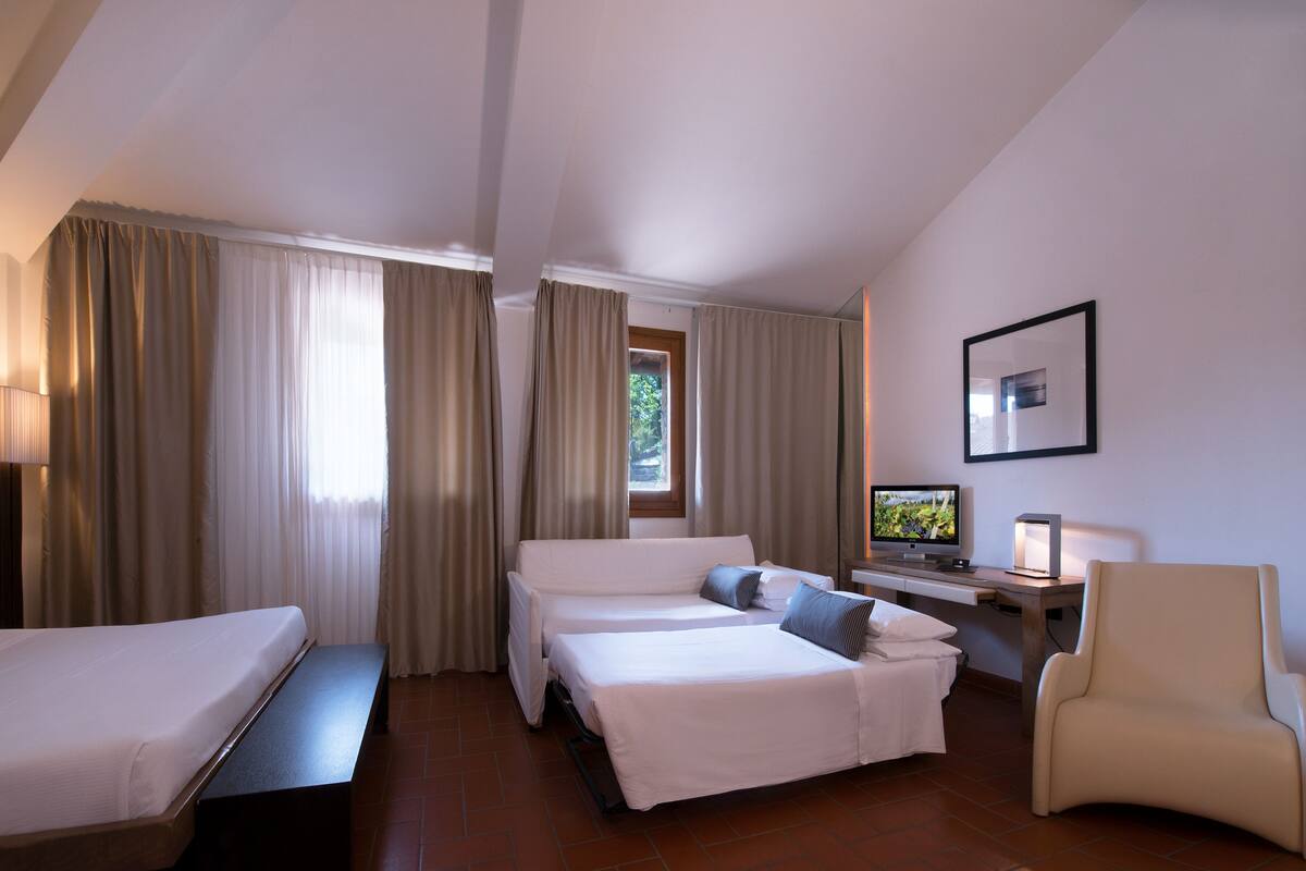 Ein Hotelzimmer mit zwei Betten, einem Schreibtisch mit Computer, einem Stuhl und einem Fenster mit Vorhängen.