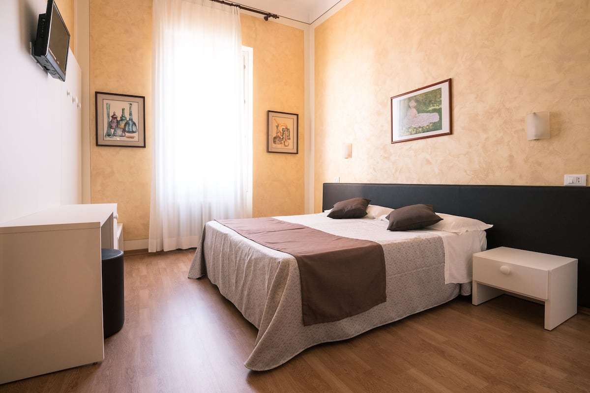 A hotel room with a bed, bedside tables, a desk, and artwork on the walls.