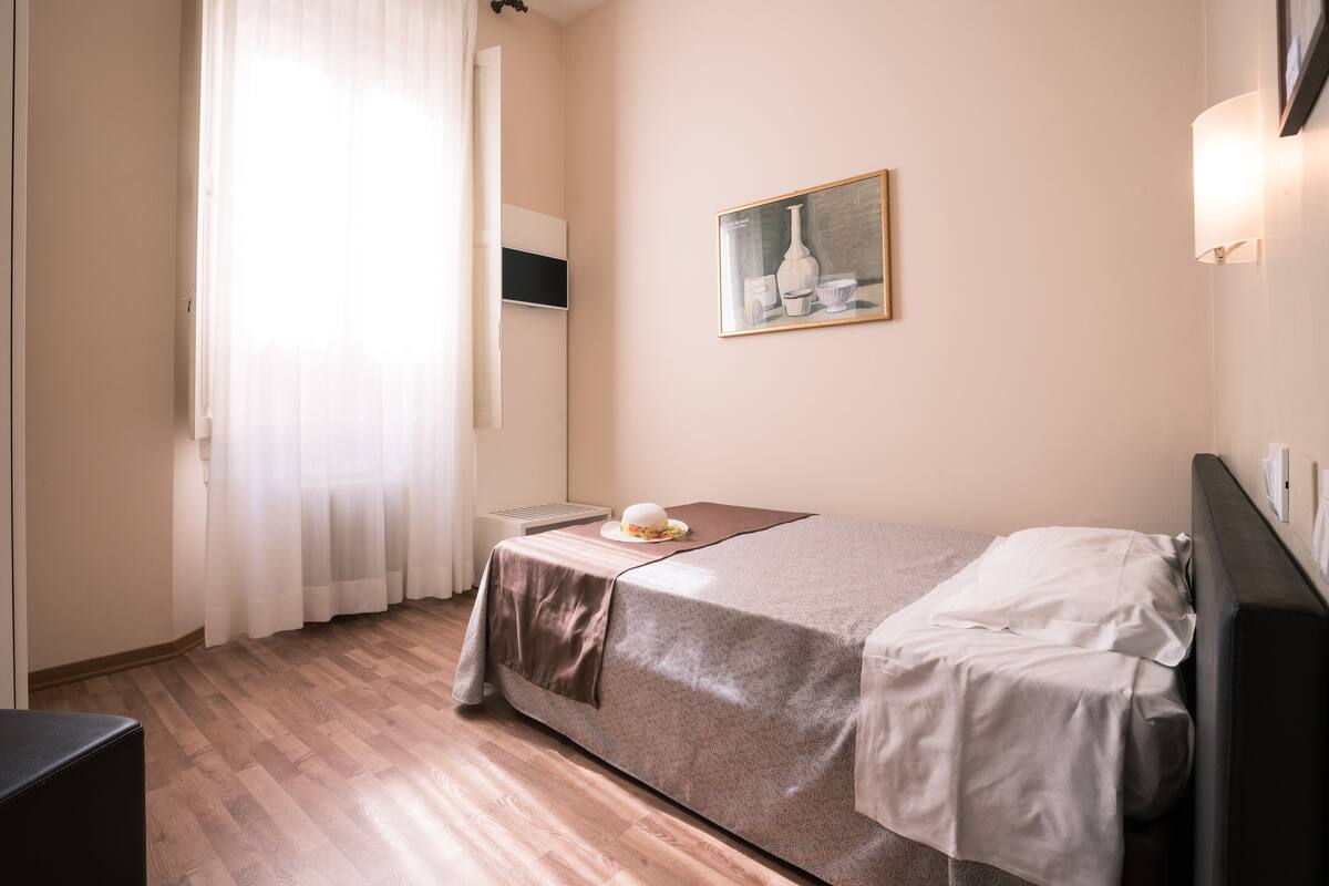 A hotel room with a bed, a bedside table, a lamp, a window with curtains, and a painting on the wall.