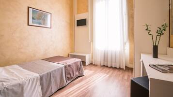 A hotel room with a bed, a desk, a chair, a potted plant, and a painting on the wall.